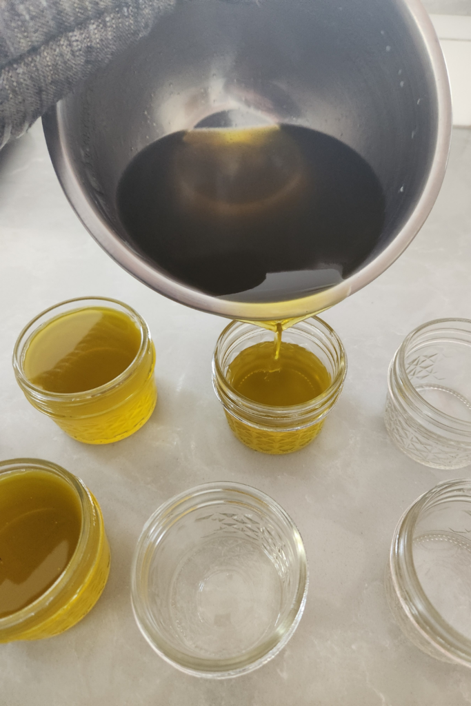 Pouring warm calendula salve mixture from a stainless steel bowl into small glass jars — golden and fragrant from shea butter and beeswax.