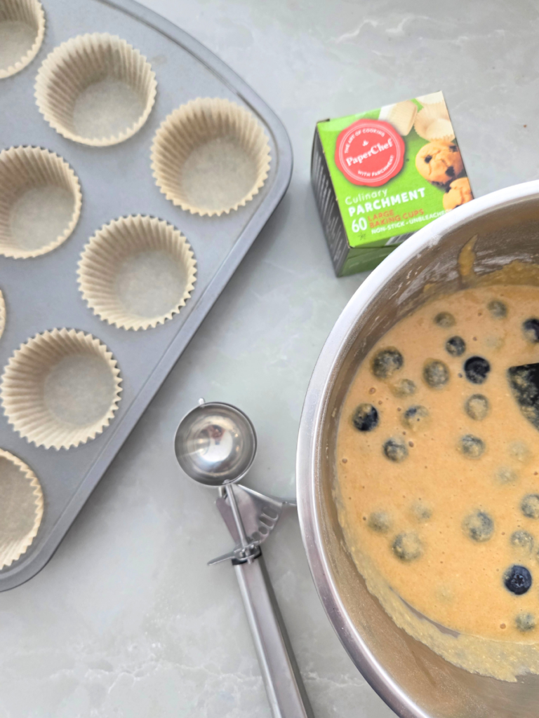 Muffin tin lined with paper liners next to a bowl of fresh milled blueberry muffin batter.