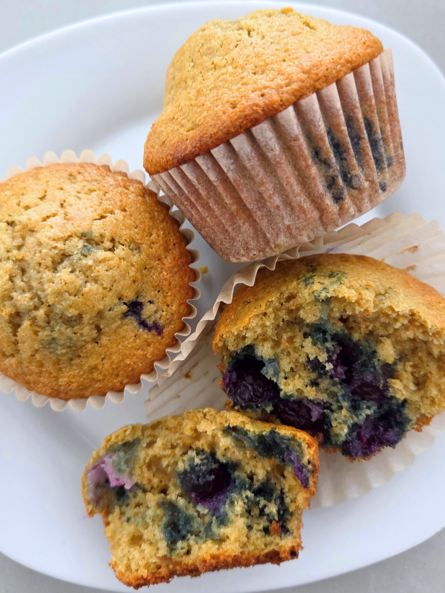 Fresh milled flour blueberry muffins on a white plate, showing golden tops and a cut muffin with a moist crumb and baked-in blueberries.