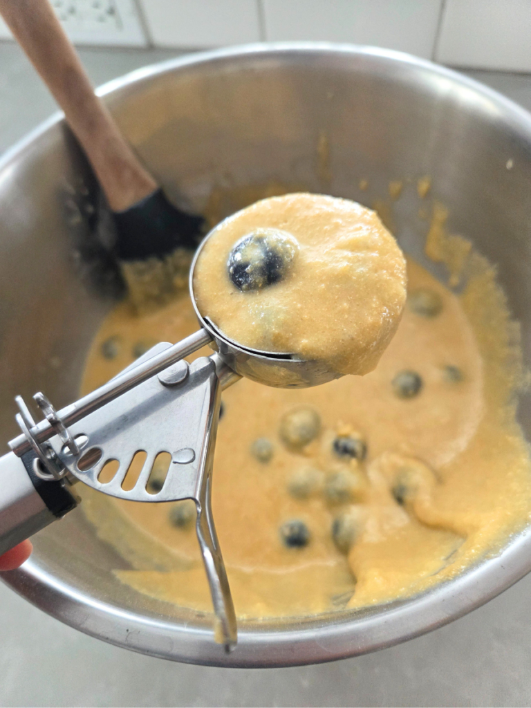 Blueberry muffin batter being scooped with a cookie scoop into prepared muffin liners.