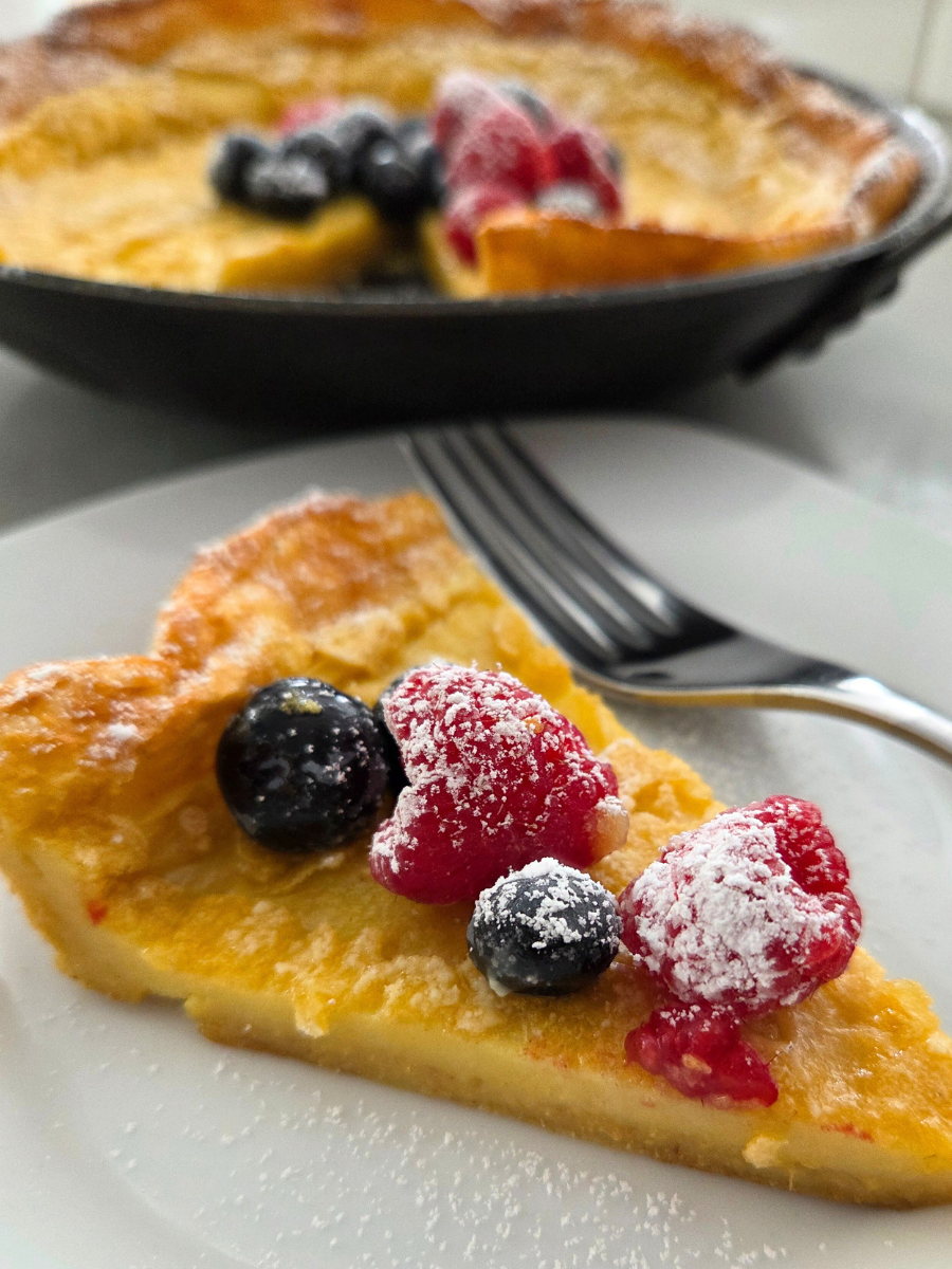 Slice of fresh milled flour Dutch baby pancake topped with berries and powdered sugar on a plate
