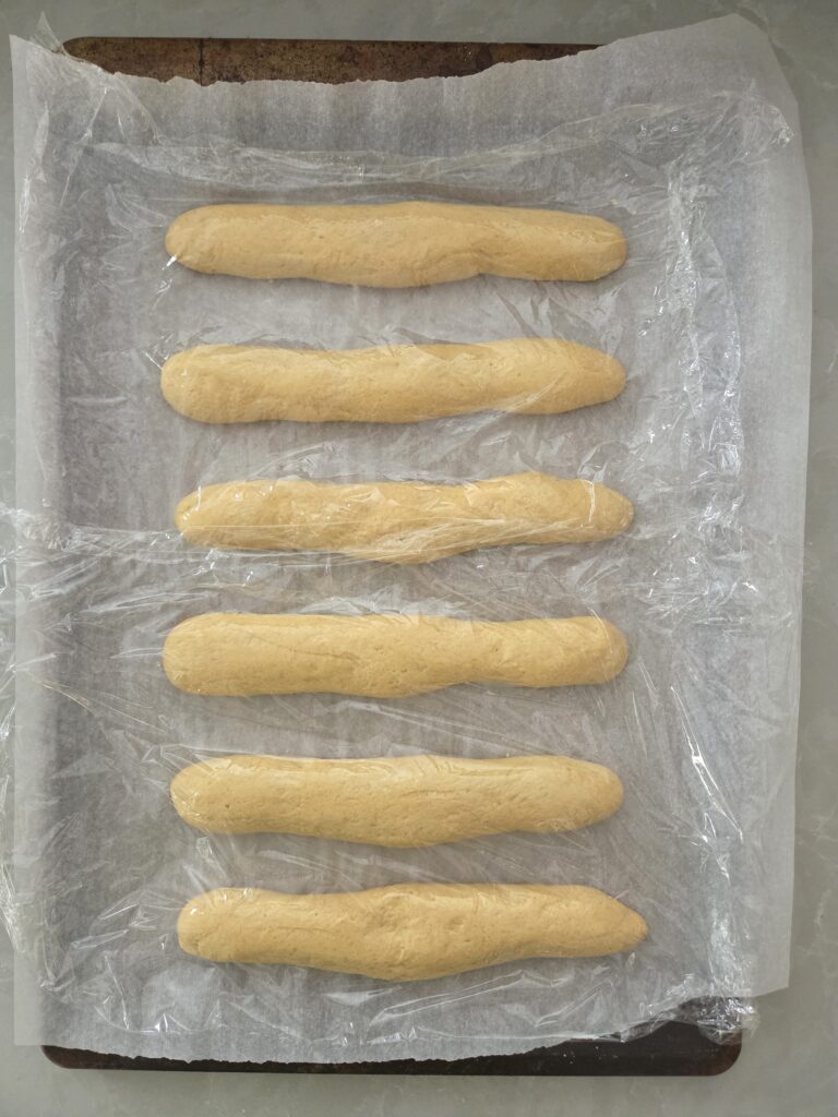 Fresh milled breadstick dough shaped and placed on a parchment-lined baking sheet, covered and ready for second rise.