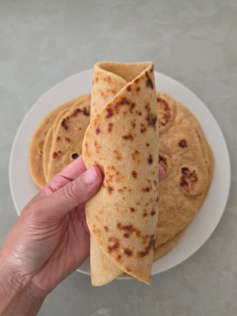 Hand holding a folded piece of homemade fresh milled flour flatbread to demonstrate its soft, pliable texture.