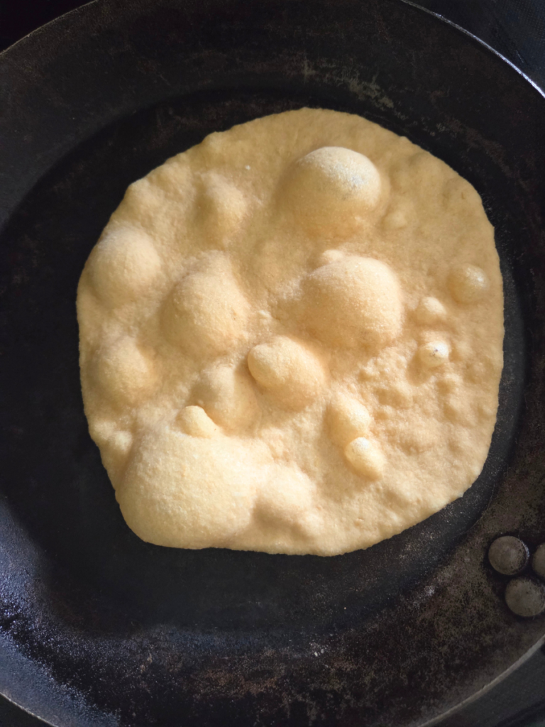 Flatbread cooking in a hot skillet with large bubbles forming across the surface.