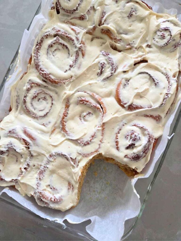 Homemade freshly milled cinnamon rolls with cream cheese frosting after one roll has been removed from the baking dish.