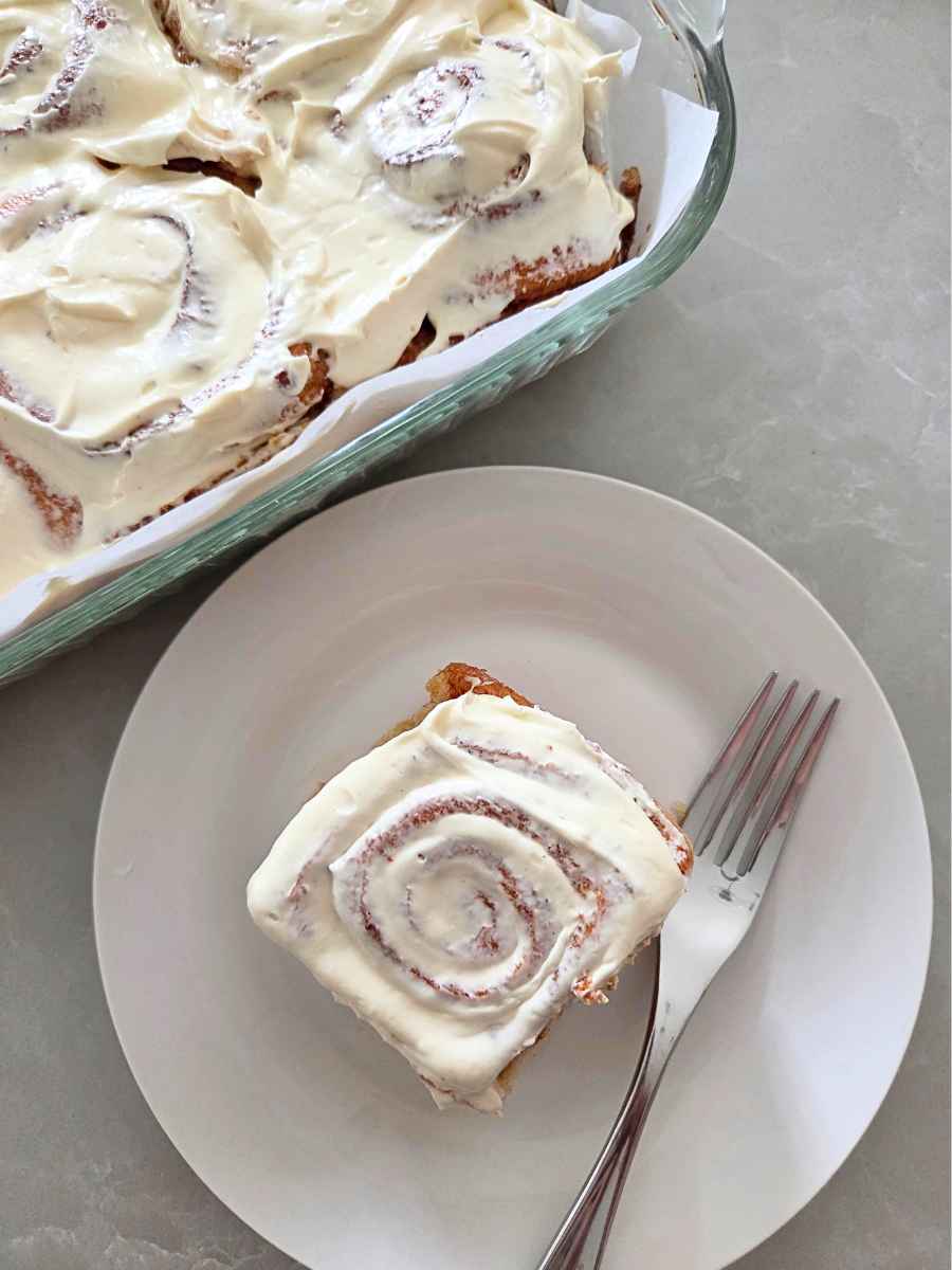 Fresh milled flour cinnamon roll with cream cheese frosting on a plate beside the baking dish.