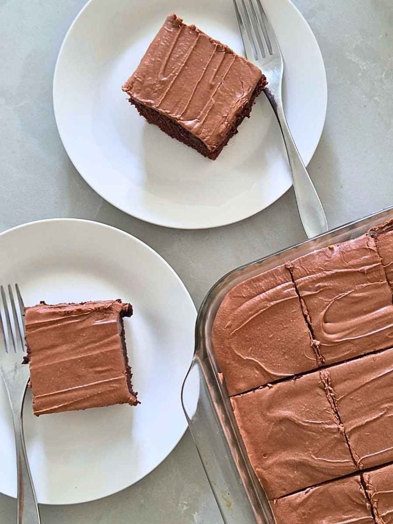 Overhead view of fresh milled flour chocolate zucchini cake cut into squares with two slices served on white plates.