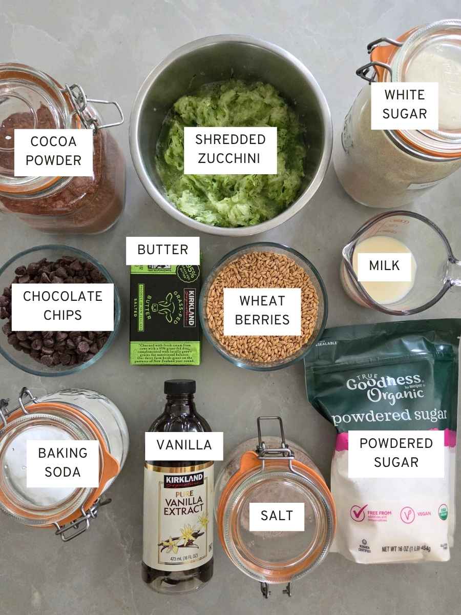 Ingredients for fresh milled chocolate zucchini cake, including wheat berries, shredded zucchini, cocoa powder, chocolate chips, butter, sugar, milk, and baking ingredients.