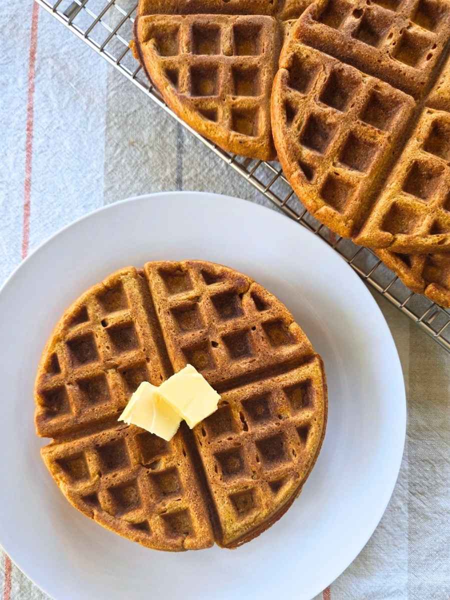Fresh milled flour pumpkin waffle topped with butter with more waffles cooling on a rack