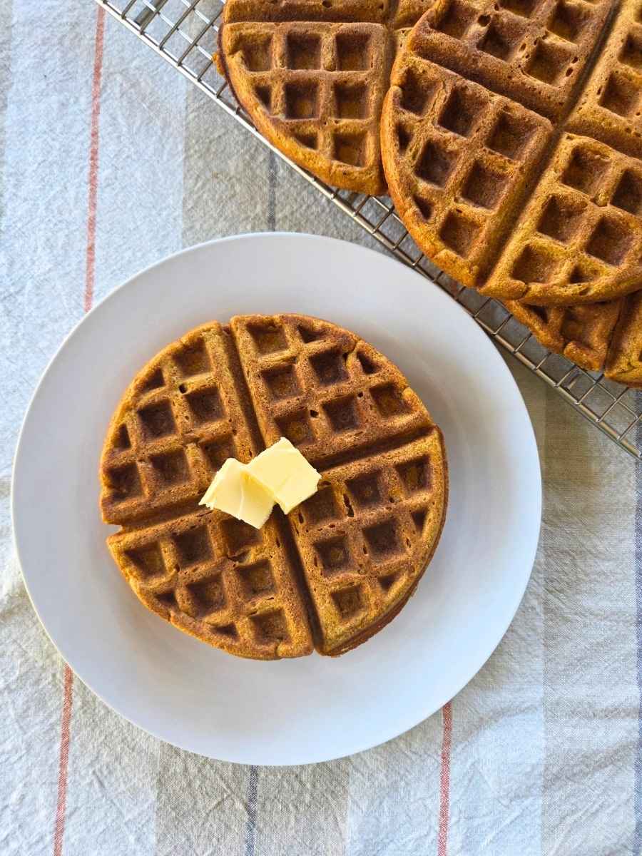Fresh milled flour pumpkin waffle topped with butter with more waffles cooling on a rack