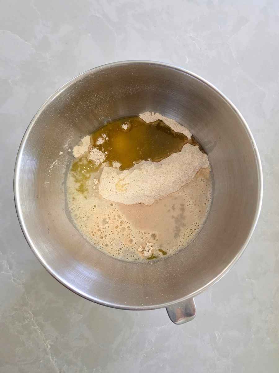 Whole wheat flour, water, olive oil, and other bread ingredients added to a stand mixer bowl before mixing.
