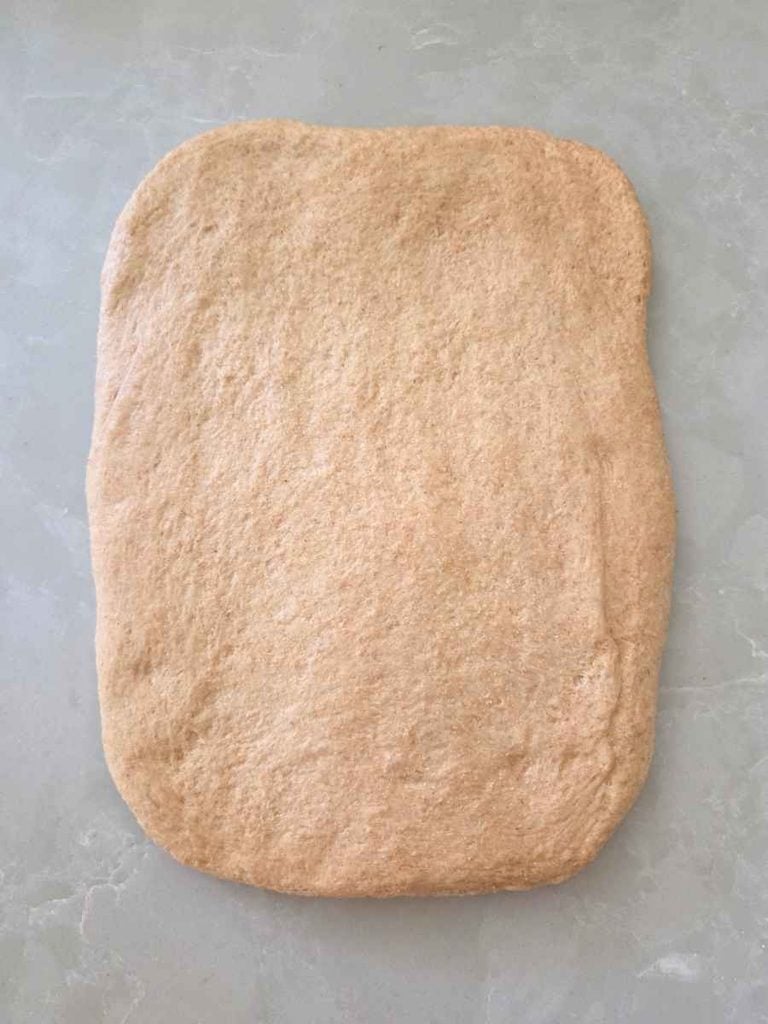 Whole wheat bread dough flattened into a rectangular shape on a countertop, ready to be rolled into a loaf.