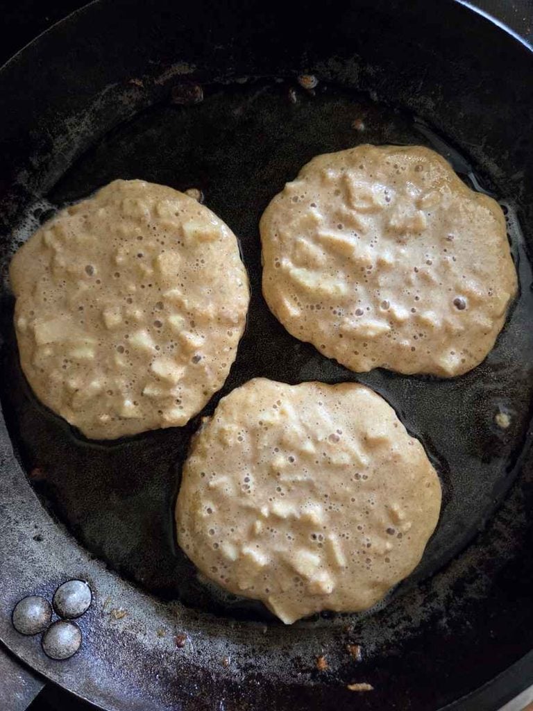 Three apple-filled pancakes cooking in a cast iron skillet with bubbles forming on the surface.