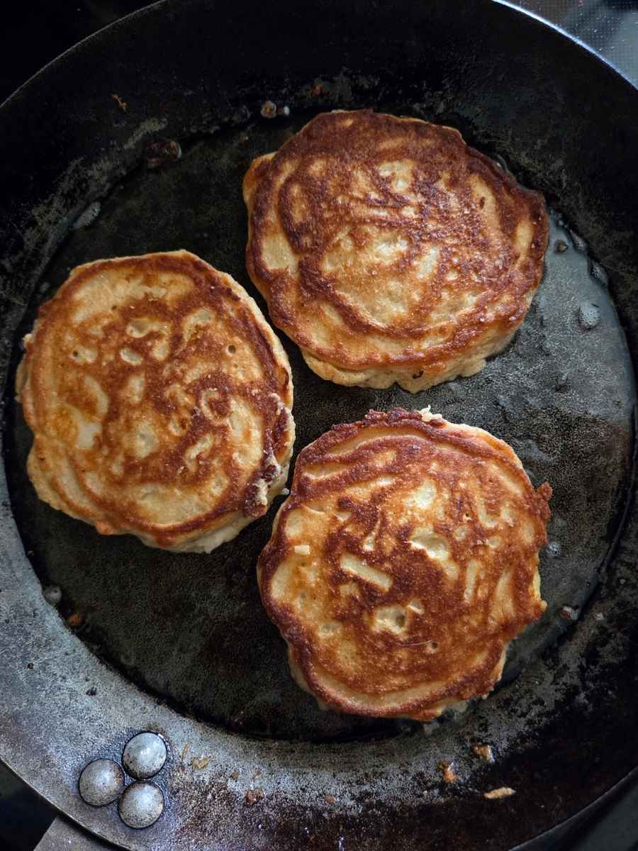 Three golden brown apple fritter pancakes with crispy edges cooking in a cast iron skillet.