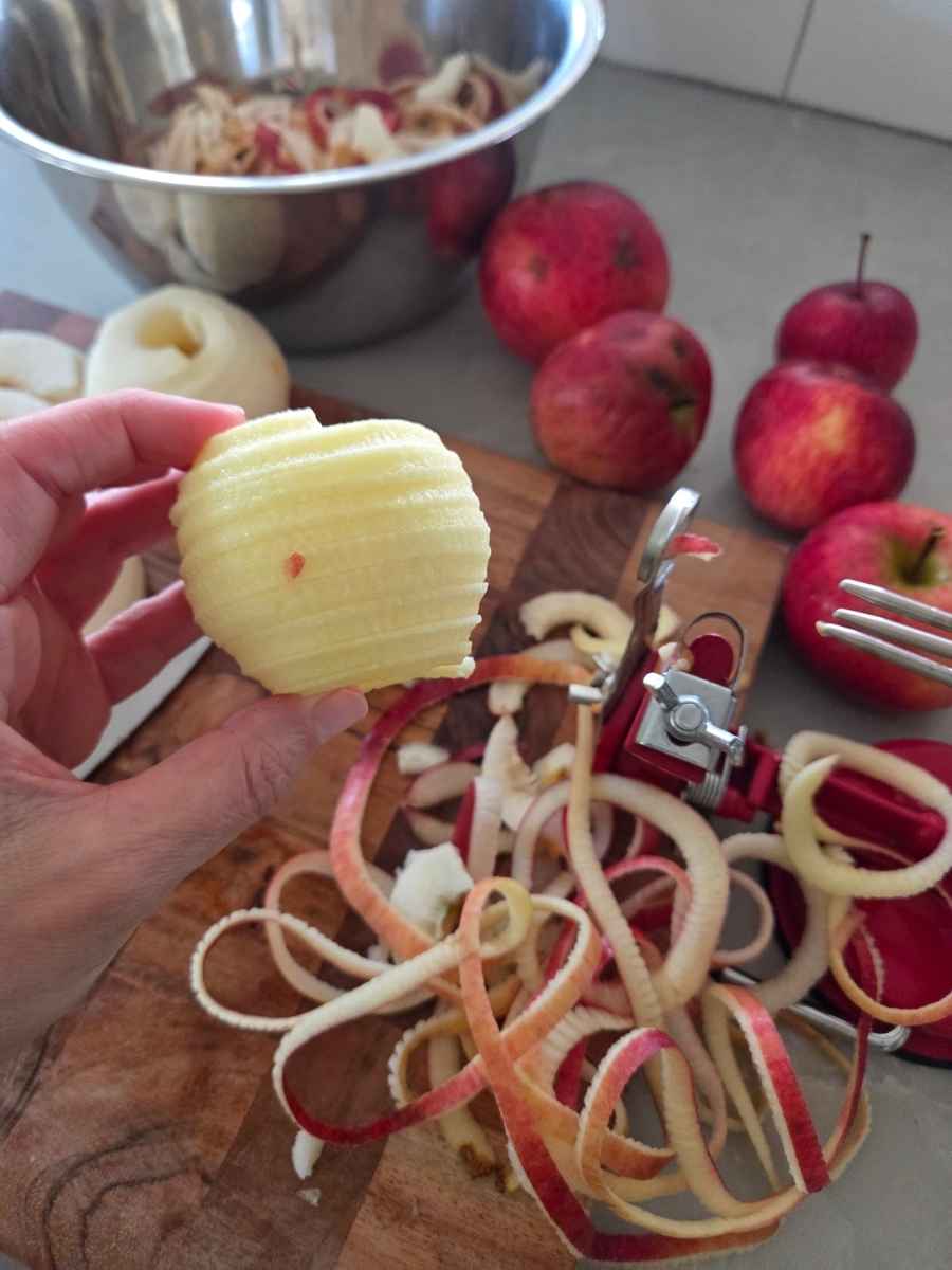 Peeled, cored, and sliced apple held beside an apple peeler/corer/slicer with apple peels on a cutting board.