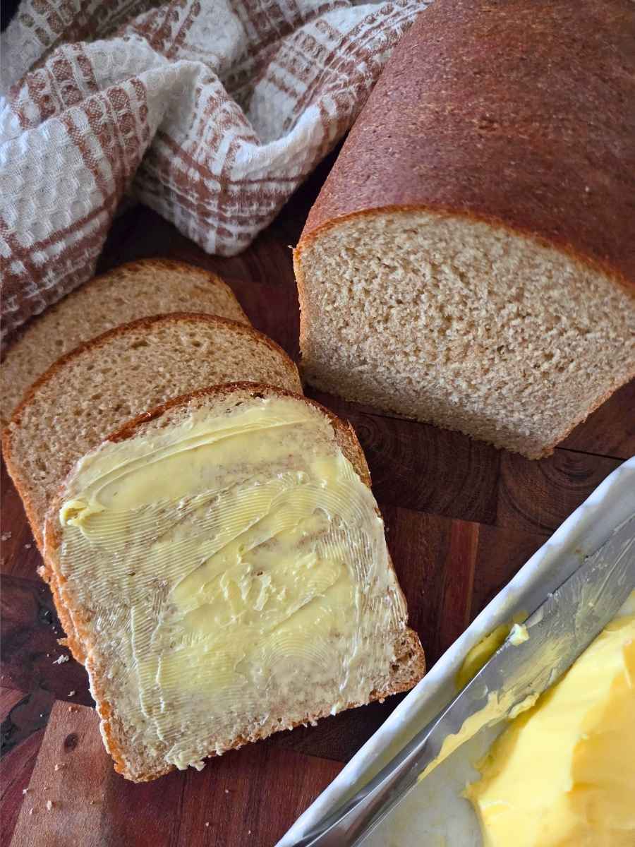 Slice of homemade fresh milled flour sandwich bread spread with butter, with additional slices and a freshly baked loaf on a wooden cutting board.